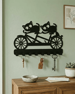 A black metal wall-mounted key rack shaped like a tandem bicycle with two cats riding, shown hanging on a pale green wall above a wooden console table with keys and decor.