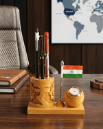 A wooden desk organizer set on a dark wood desk, featuring a carved cylindrical pen holder with three pens, a small Indian flag on a stand, and a round wooden clock.