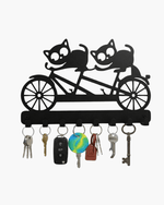 Black metal key holder shaped like two cats riding a tandem bicycle, featuring 8 hooks holding various keys on a white background.