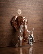 A copper elephant head key holder mounted on a dark wooden textured wall. A keyring with old-fashioned keys hangs from the left white tusk, and a modern car key fob hangs from the right tusk. A small round logo for