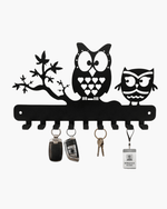 Black metal owl wall key holder mounted on a textured gray wall above a wooden console table. The holder features two owls, a tree branch motif, and multiple key hooks holding keys, a car key fob, and an ID badge.