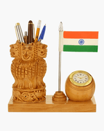 A wooden desk decor set featuring a carved Lion Capital of India, a small Indian flag on a silver pole, and a round wooden clock with a gold-tone bezel, all mounted on a rectangular wooden base against a light gray background.