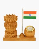 A wooden desk decor set featuring a carved Lion Capital of India, a small Indian flag on a silver pole, and a round wooden clock with a gold-tone bezel, all mounted on a rectangular wooden base against a light gray background.