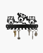 Black metal owl wall key holder with three owl silhouettes on a branch and several keys hanging from hooks, shown on a plain light background.