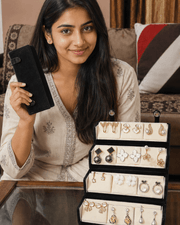 A woman holds a black velvet jewelry box while wearing a beige embroidered kurta. In front of her is a tiered black jewelry organizer filled with gold, silver, and white stud earrings and necklaces.