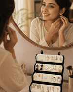 A woman looks into a round mirror while trying on a gold earring, with her reflection visible. A black jewelry organizer filled with assorted stud earrings sits on the counter below.