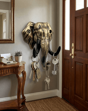 An antique gold elephant head key holder mounted on a gray wall in a home entryway next to a wooden door. Sets of keys hang from its tusks and trunk. A wooden console table sits to the left