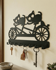 Close-up wall-mounted view of decorative black metal cats on bicycle key hanger with keys hanging on standard entryway wall.