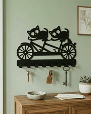 A black metal wall-mounted key rack shaped like a tandem bicycle with two cats riding, shown hanging on a pale green wall above a wooden console table with keys and decor.
