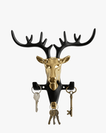 Golden deer head wall hook holding multiple sets of keys and a vintage brass key against a white background