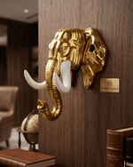 A glossy gold elephant head wall hook mounted on a dark wood panel. A set of keys hangs from one of its white tusks, and a vintage key hangs from the curled trunk. A small gold placard reading handicrush.com is mounted to the right