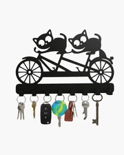 Black metal key holder shaped like two cats riding a tandem bicycle, featuring 8 hooks holding various keys on a white background.