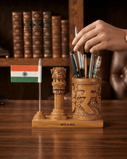 A carved wooden desk organizer set on a wooden table, featuring a small Indian flag, a lion capital pillar sculpture, and a cylindrical holder carved with the Indian Rupee symbol and filled with pens. A hand is placing pens into the holder.