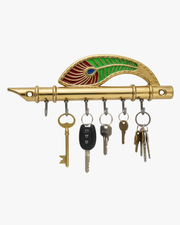 A decorative gold-toned brass key holder shaped like a traditional Indian flute (Bansuri), featuring an ornate enameled peacock feather in green, red, and blue. The holder includes six sturdy metal hooks and measures 7 inches in length and 3 inches in height
