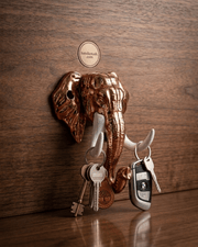 A copper elephant head key holder mounted on a dark wooden textured wall. A keyring with old-fashioned keys hangs from the left white tusk, and a modern car key fob hangs from the right tusk. A small round logo for 