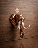 The copper-finished elephant head key hanger with white tusks mounted on a dark wooden wall without any keys hanging from it. A circular logo for 