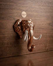 The copper-finished elephant head key hanger with white tusks mounted on a dark wooden wall without any keys hanging from it. A circular logo for 