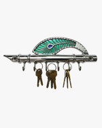 A wall-mounted silver metal key holder shaped like a flute with a green peacock feather design, shown with three sets of keys hanging from the hooks