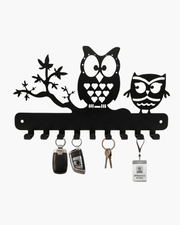 Black metal owl wall key holder mounted on a textured gray wall above a wooden console table. The holder features two owls, a tree branch motif, and multiple key hooks holding keys, a car key fob, and an ID badge.