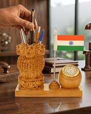 A carved wooden pen holder shaped like the Ashoka Chakra sits on a wooden desk base next to a small Indian flag and a round wooden clock. A hand is placing pens into the holder, with books and a lamp in the background.